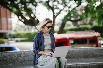 A cheerful young woman sits at a city park working with her laptop, combining work and leisure,...