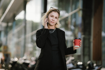 A young woman enjoys coffee outdoors while browsing her smartphone, embracing a modern urban...