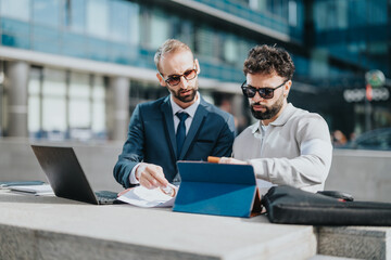 Two business people collaborate outdoors, using laptops and tablets for discussing work. Their modern yet professional interaction reflects productivity, teamwork, and technology in a business area.