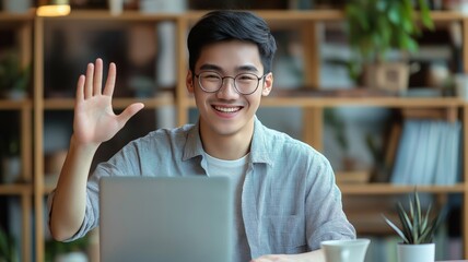 Smiling young man waving hello while using a laptop in a cozy indoor setting with plants