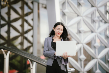 Fototapeta premium Confident, determined businesswoman Standing in front of a modern office building holding a laptop, showing ambition, leadership and empowerment in today’s fast-changing digital and corporate world.