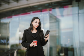 A confident, stylish woman in a black blazer walks through the city with coffee and smartphone in...