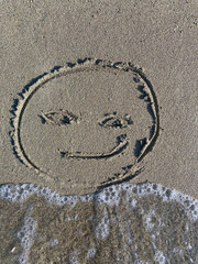 Playful smiley face drawn in the sand along a sunny beach against gentle ocean waves