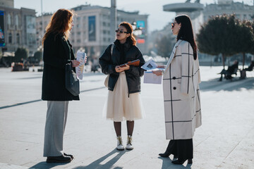 Three women are seen standing outdoors in a city square, discussing and sharing documents. The...