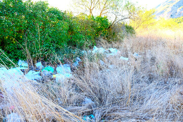 Discarded plastic bags and rubbish litter a dry grassy area with green bushes in the background, highlighting environmental pollution.