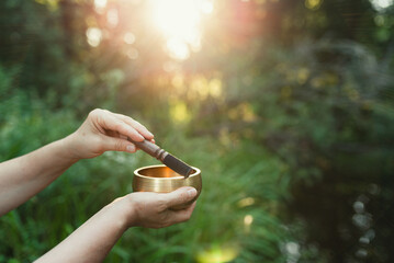 Close-up of a woman holding a Tibetan singing bowl outdoors, with soft sunlight filtering through blurred forest trees in the background.