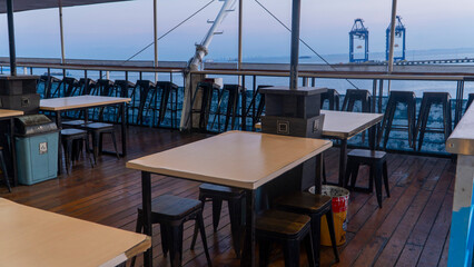 Outdoor dining area on a passenger ship deck with tables, chairs, and harbor cranes in the background