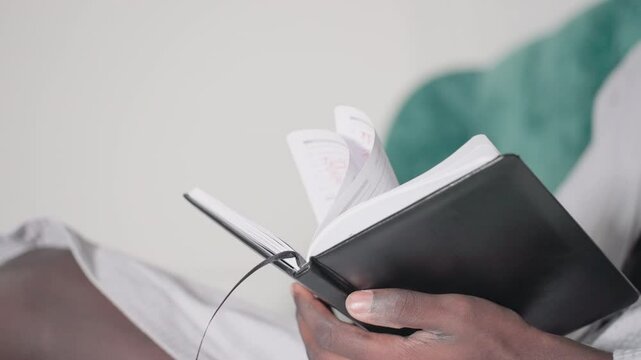 Close-up of man in white bathrobe holding open black notebook with pages flipping, soft light highlighting fingers and pages, green velvet headboard in background, peaceful indoor atmosphere