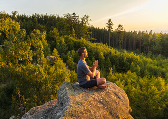 Young man meditating, praying on top of rock with trees behind. Active lifestyle, mental health...