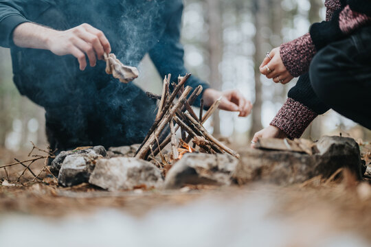 A close-up photograph depicting two individuals collaborating to construct a campfire amidst an outdoor natural setting, conveying themes of wilderness, survival, and companionship.