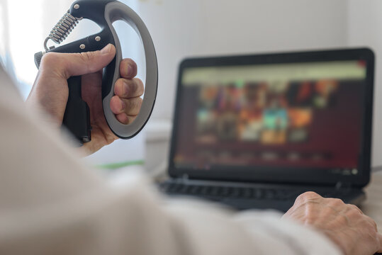 Closeup photo of man queezing expander in hand and looking at laptop screen. Close-up of male hand exercising with grip trainer near laptop