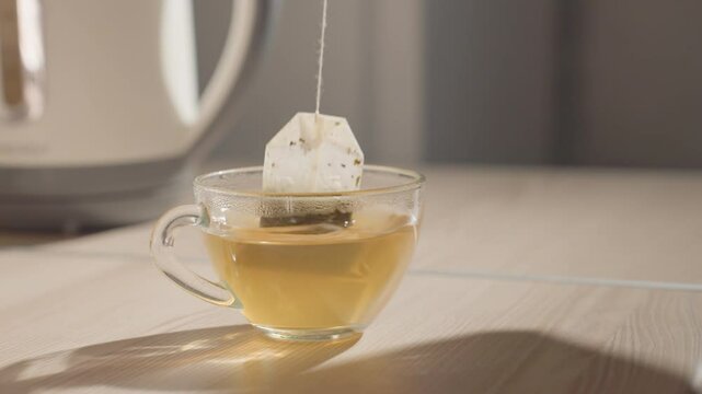 Hand reaching into a cup with tea bag floating inside, pouring hot water from kettle. Close-up of cup and tea bag steeping in hot liquid on kitchen table, preparing drink in calm morning setting