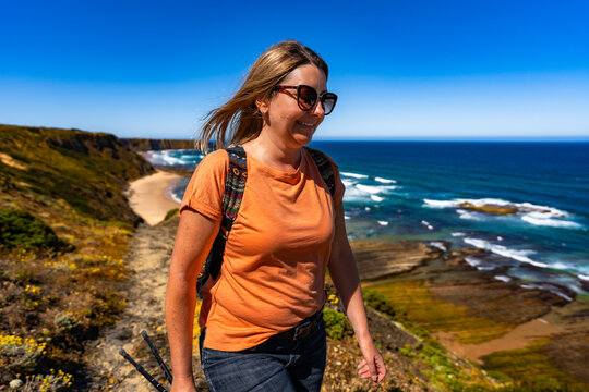 Beautiful mid adult woman tourist hiking on trail along Atlantic ocean in nature park Vicentine Coast in Algarve in Portugal on summer day. Side view	 - Powered by Adobe