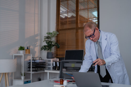 Experienced senior doctor in a white coat and glasses analyzing data on a laptop in a modern office setting with natural light and organized workspace elements