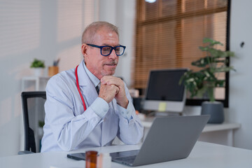 Experienced senior doctor wearing glasses conducting an online consultation from a modern office. The workspace is equipped with a laptop and contemporary decor, enhancing professionalism