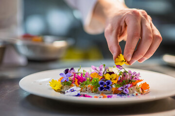 Mid-action shot of chef’s hand placing edible flowers on artistically arranged fine dining plate, stainless steel kitchen setting