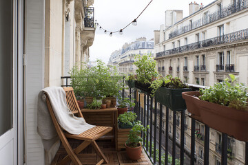 Tight view of a narrow city balcony with herb pots, folded blanket on chair, and string lights above, early morning