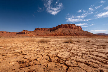 Wide shot of arid desert terrain with cracked ground texture, distant red mesa under bright blue sky
