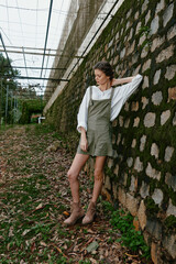 Young woman in casual summer outfit stands against a moss covered wall, showcasing a relaxed vibe...