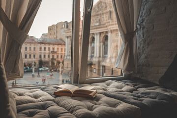 Cozy reading nook with an open book on a cushioned window seat overlooking a historic European city...