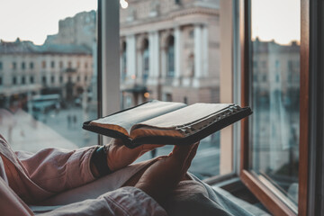 Person reading a Bible by the window with a view of the Lviv Opera House in the background during...
