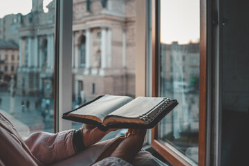 Person reading a Bible by the window with a view of the Lviv Opera House in the background during...
