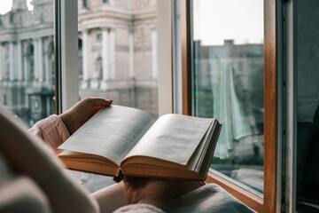 Person holding an open vintage book by a window with a view of a historic European square and opera...