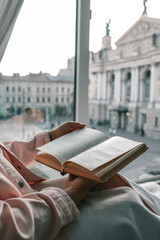 Person holding an open vintage book by a window with a view of a historic European square and opera...