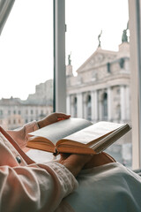 Person holding an open vintage book by a window with a view of a historic European square and opera...