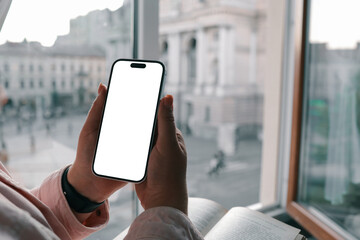 Woman holding smartphone with blank screen while reading a book by the window with blurred view of Lviv Opera House in the background. Cozy, soft lighting and thoughtful atmosphere