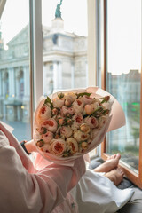 Woman holding a bouquet of delicate pastel roses by an open window with a view of Lviv Opera House....