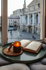 Open book and lit candle on a cozy windowsill nook with cushions, overlooking the Lviv Opera House....
