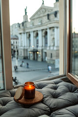 Burning candle in amber glass jar with wooden lid on a cork coaster, placed on a soft grey cushion...