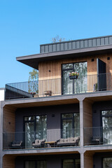 A contemporary building with wooden paneling, black railings, furnished balconies, and large glass doors. Trees are partially visible above.