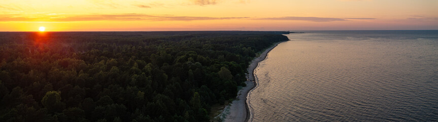 Sunset over a coastal area with a dense forest along the shoreline, a narrow sandy beach, and calm...