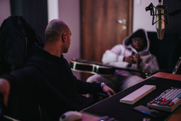 A dynamic image of two musicians in a recording studio, one holding a guitar, and another turning towards him. A microphone is prominent, suggesting a collaborative music creation session.