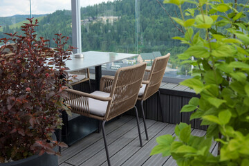 Outdoor terrace with modern table and wicker chairs surrounded by green plants. Scenic mountain and...