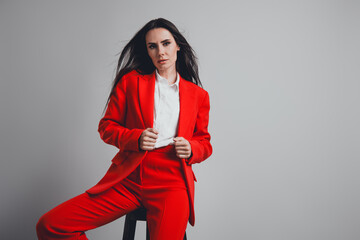 Confident young professional woman in elegant red suit posing against grey background, showcasing...
