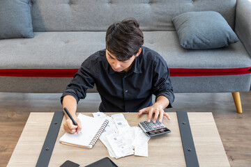 Young Asian man calculating expense budget bills finance using calculator sitting on floor in...