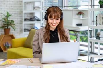 Young Asian woman wearing headphones working on laptop at creative desk with color swatches and sketches in modern office