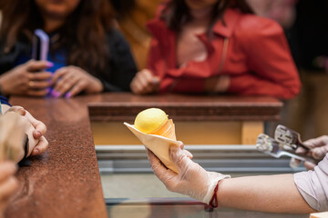 Ice cream in waffle cone. Vendor in gloves handing yellow ice cream cone to customer over counter...