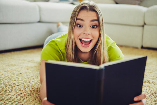 Excited young woman reading a book on a comfortable carpeted floor at home, showcasing leisure and joy in a relaxed setting