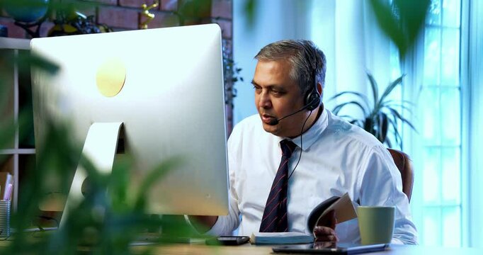Indian call center male executive taking notes while sitting at desk in modern office with headphones, Asian corporate employee attending customer service calls on computer system with focus