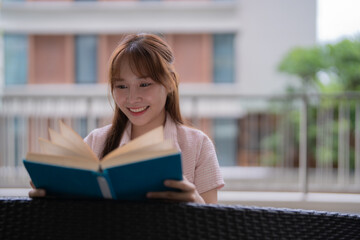 Young woman with long brown hair, wearing a pink shirt, sitting on a balcony and smiling while enjoying a blue book, embracing a moment of relaxation and happiness in the sunlight