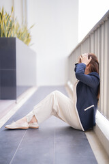 Stressed businesswoman suffering from a headache while sitting on the floor near railing, covering...