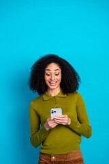 Cheerful young woman using smartphone against bright blue backdrop, expressing delight with curly...