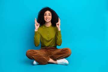 Smiling young woman with curly hair sitting on floor fingers crossed hopeful against a vibrant blue...