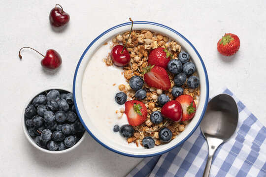 Natural yogurt with granola, blueberries and strawberries in bowl on light background with spoon and napkin. Healthy and nutritious breakfast concept. Top view and copy space.