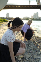 Fototapeta premium Two young women crouch by the riverside, focusing on collecting stones as the sunlight reflects off the water and city buildings in the background during late afternoon.
