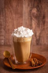 Delicious pumpkin spice latte made from coffee decorated with whipped cream topping served in drinking glass on plate with cinnamon sticks and spoon on dark brown table against rustic wooden wall